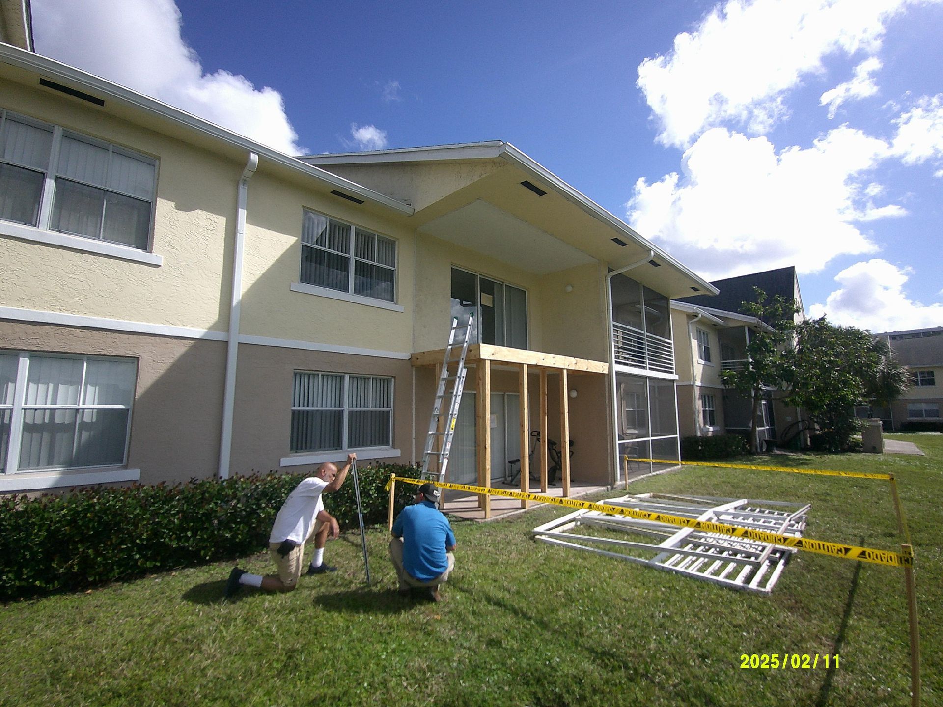Workers installing caution tape barriers on the lawn beneath a balcony under temporary wood shoring, with a removed railing frame set aside, during structural repair work in Fort Lauderdale, FL by M&P Consulting Engineers
