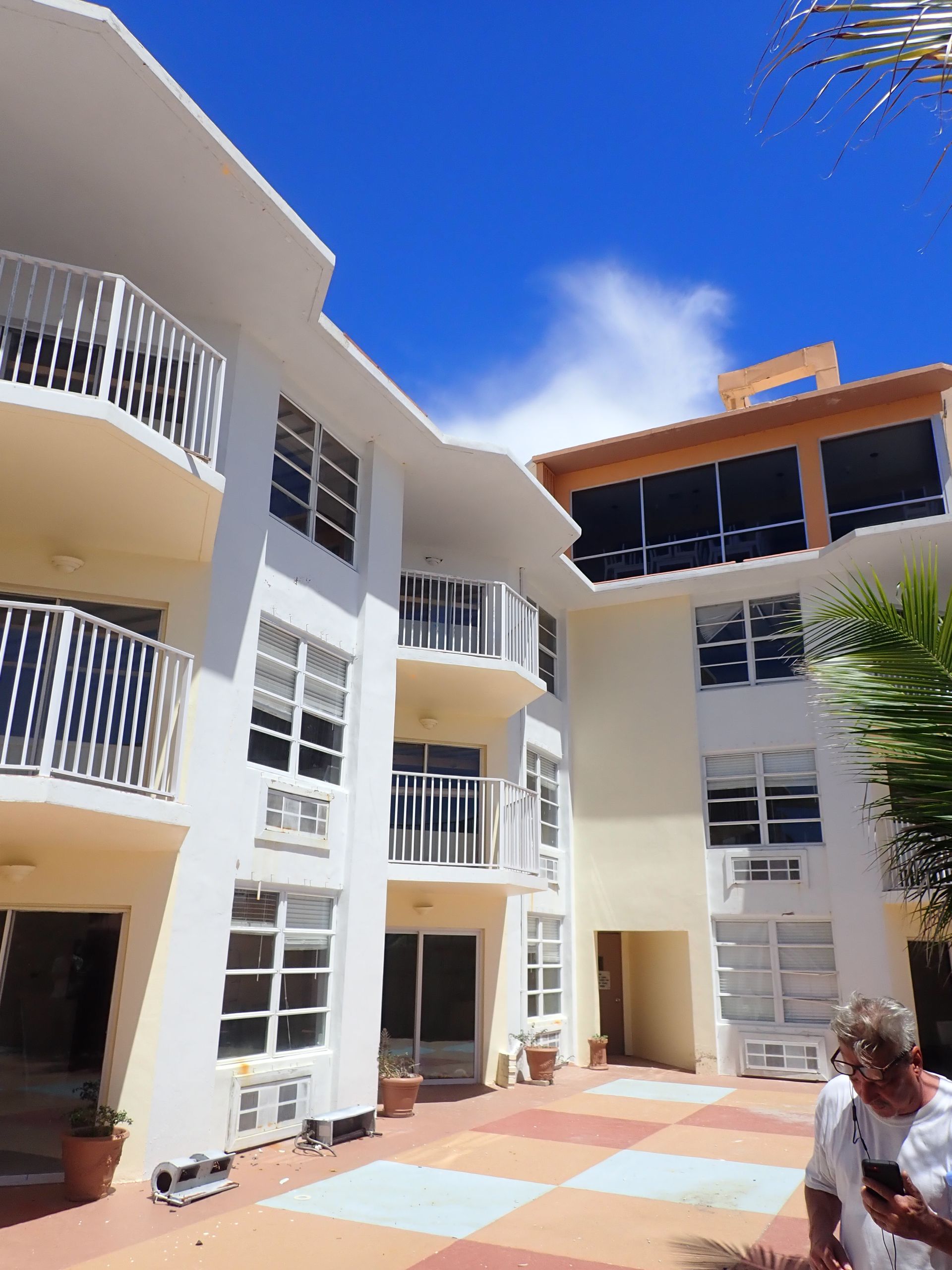 Courtyard with white balconies and windows under a bright blue sky; a person sits at lower right.
