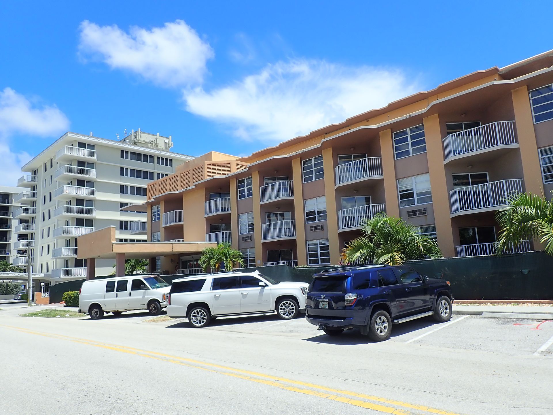 Sunny street with parked cars beside a tan apartment building under a blue sky