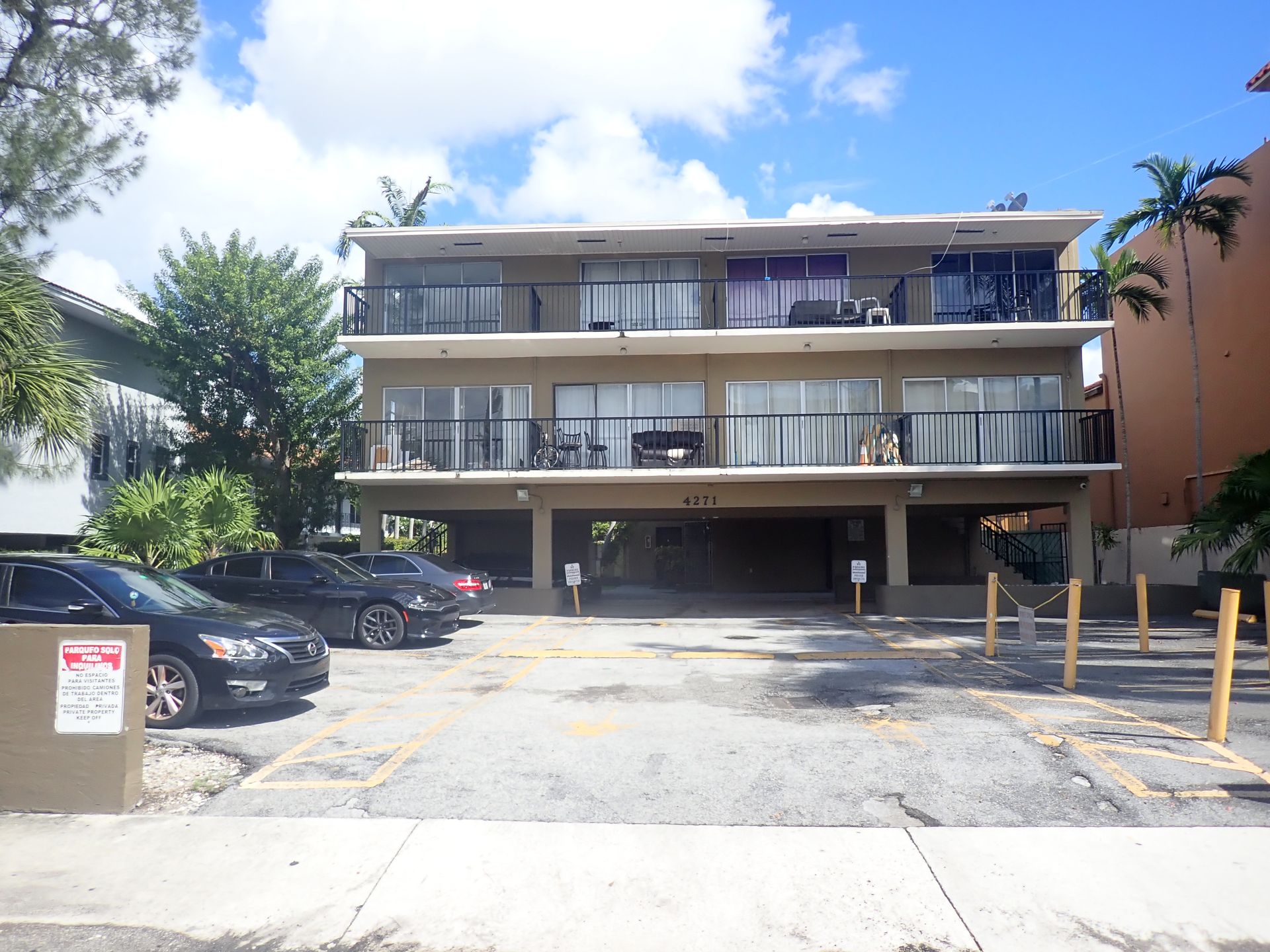 Two-story apartment building with balconies, parked cars, and a sunny parking lot.