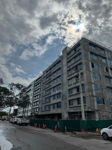 Cloudy street view of a gray apartment building behind a green construction fence, with parked cars along the road.