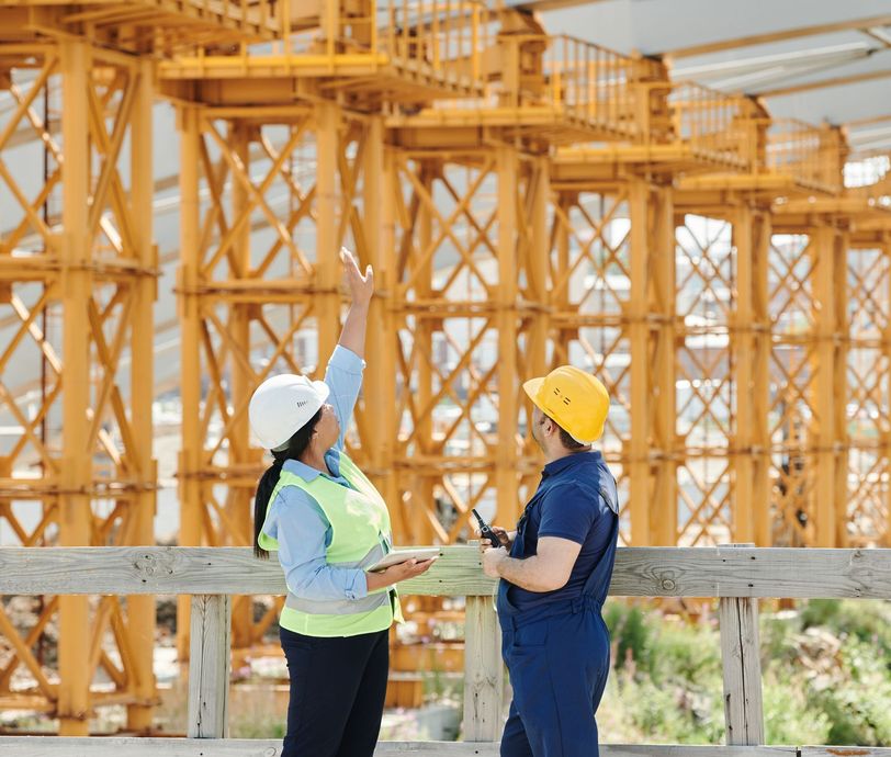 Two construction workers in hard hats pointing at a large wooden structure on site.