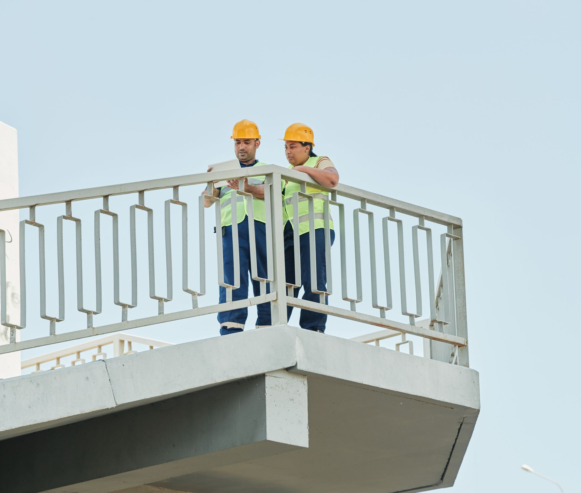 Two construction workers in hard hats stand on a metal platform against the sky.