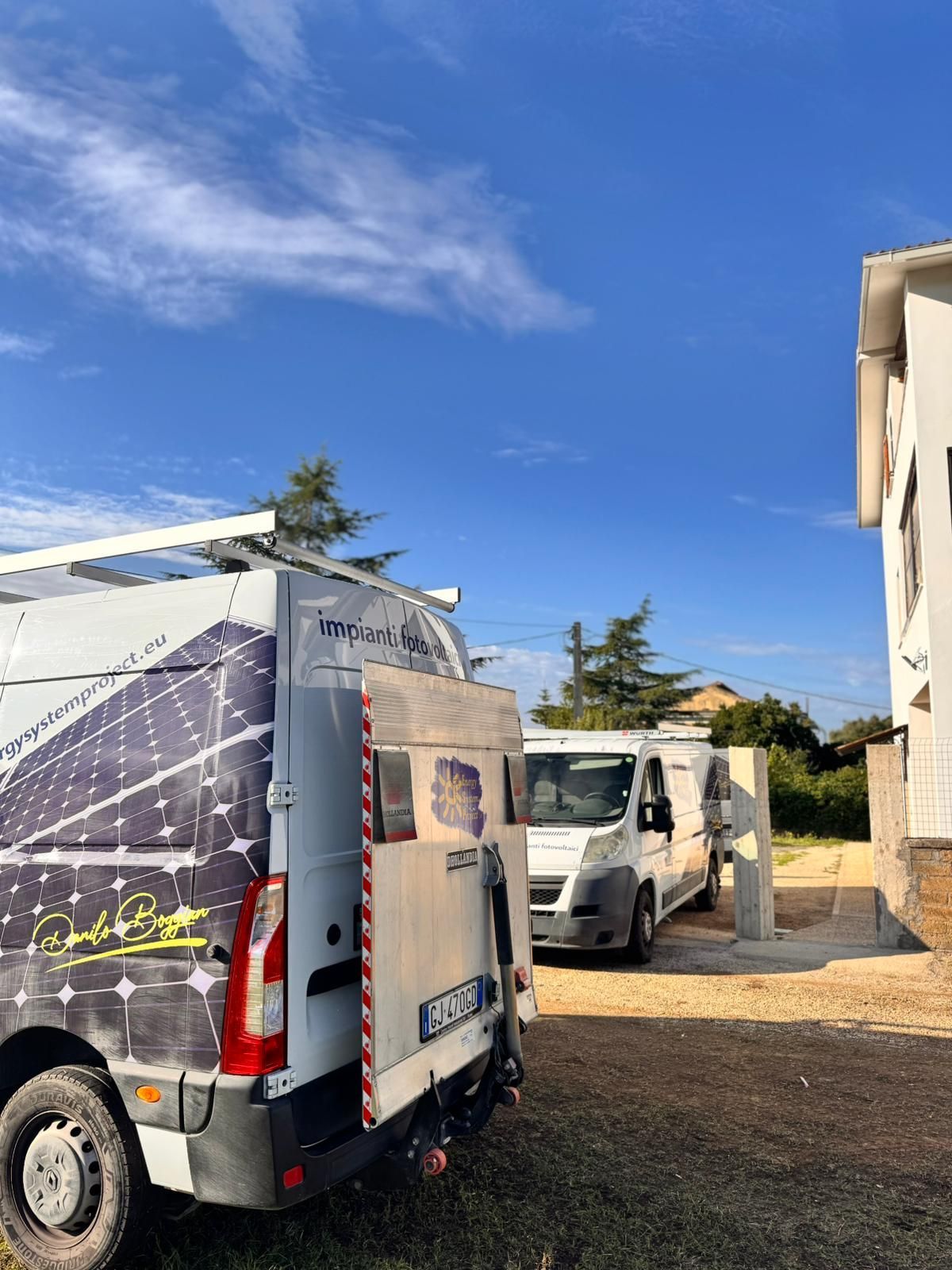 Furgone bianco parcheggiato su un piazzale ghiaioso accanto a un edificio in pietra sotto un cielo azzurro