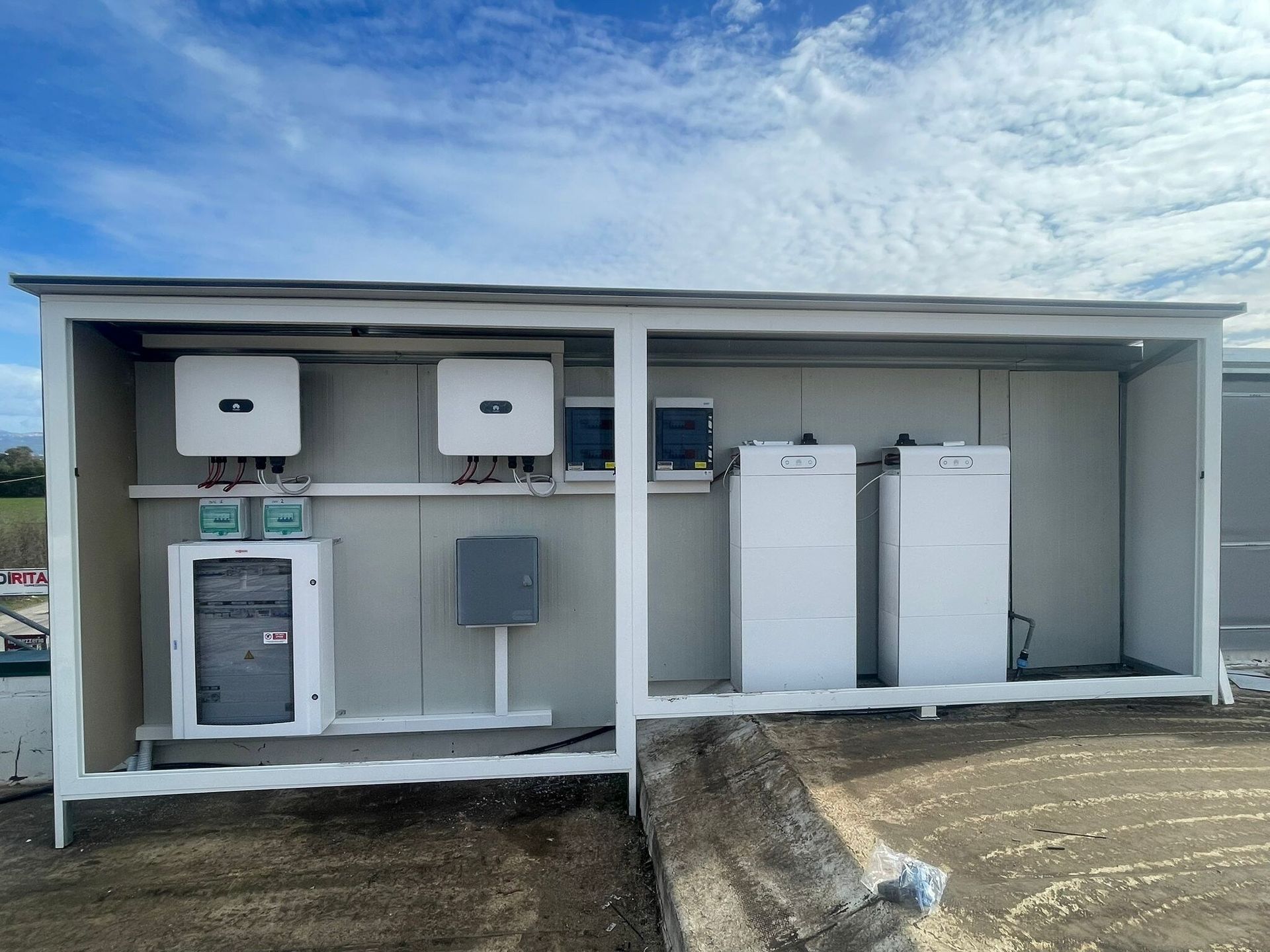 Apparecchiature elettriche esterne e quadro contatori in armadi di servizio aperti accanto a un terreno sterrato sotto un cielo azzurro.