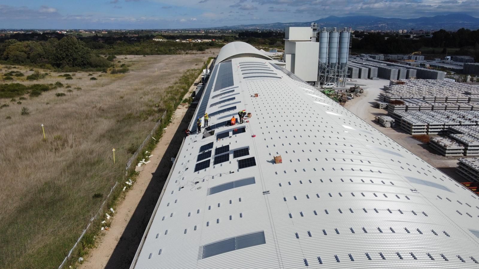 Veduta aerea di un grande tetto industriale bianco con prese d'aria, pannelli solari e un cantiere edile adiacente.