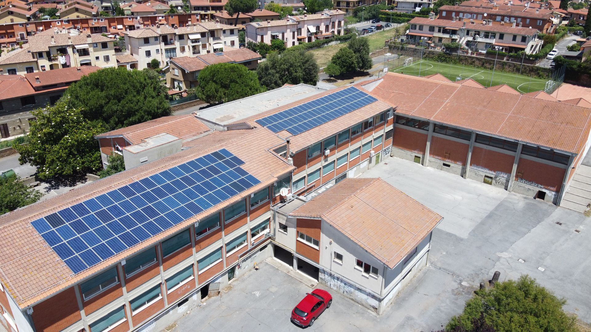 Veduta aerea di un edificio scolastico con pannelli solari sul tetto e un'auto rossa nel cortile.