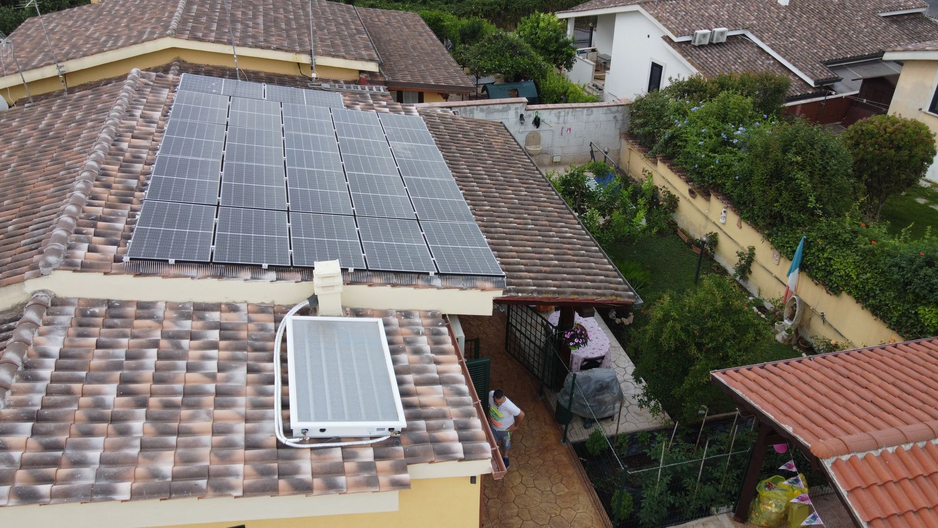 Veduta aerea del tetto di una casa con pannelli solari e lucernario, circondata da case vicine e alberi.