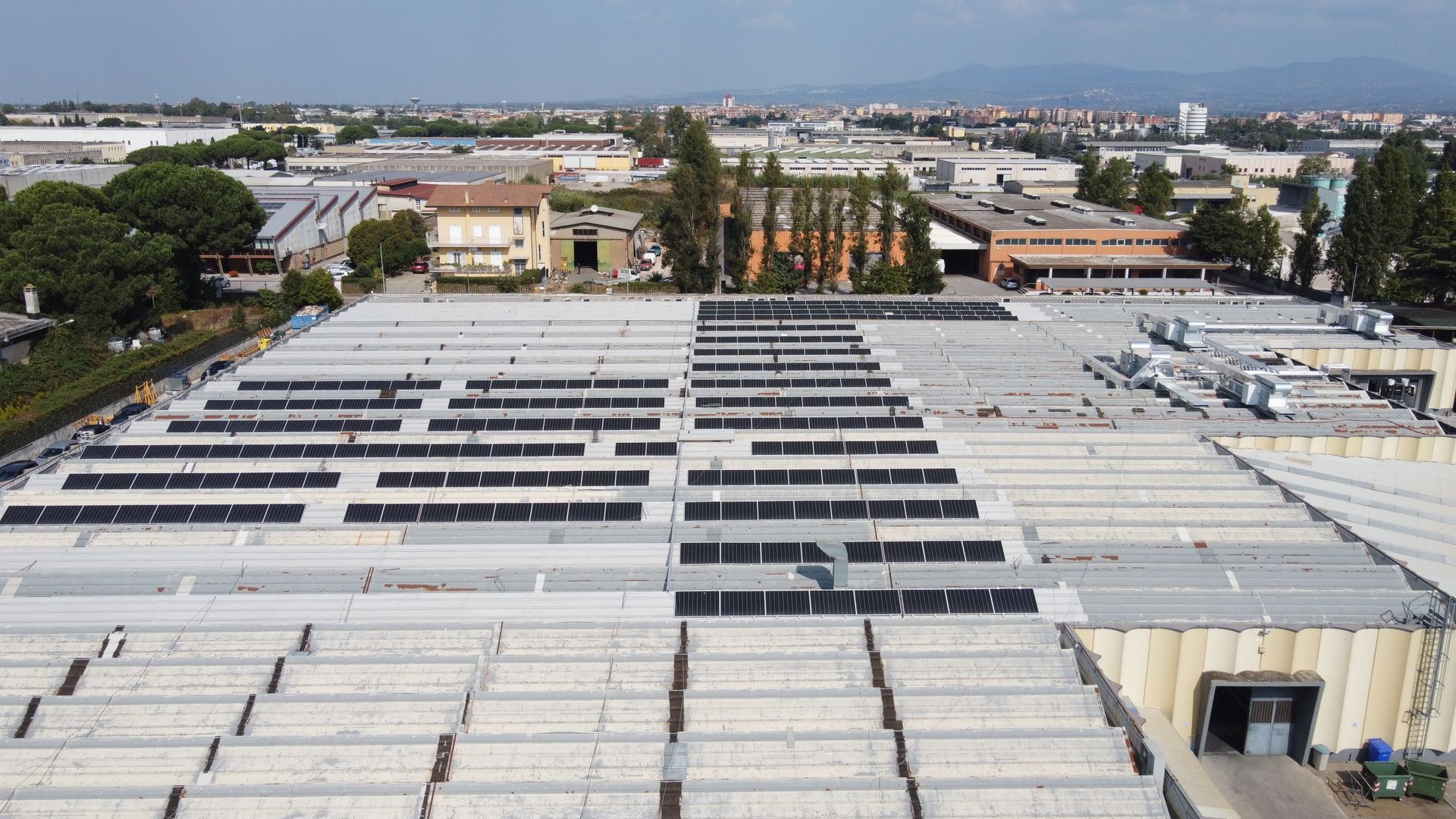 Veduta aerea di un grande tetto industriale con file di pannelli solari e degli edifici circostanti.