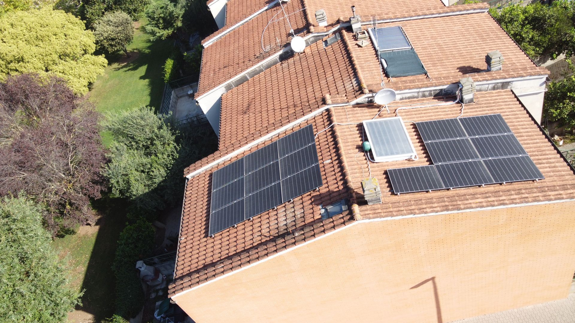 Tetto con numerosi pannelli solari su una casa con tegole in terracotta, accanto ad alberi e al tetto di un vicino.