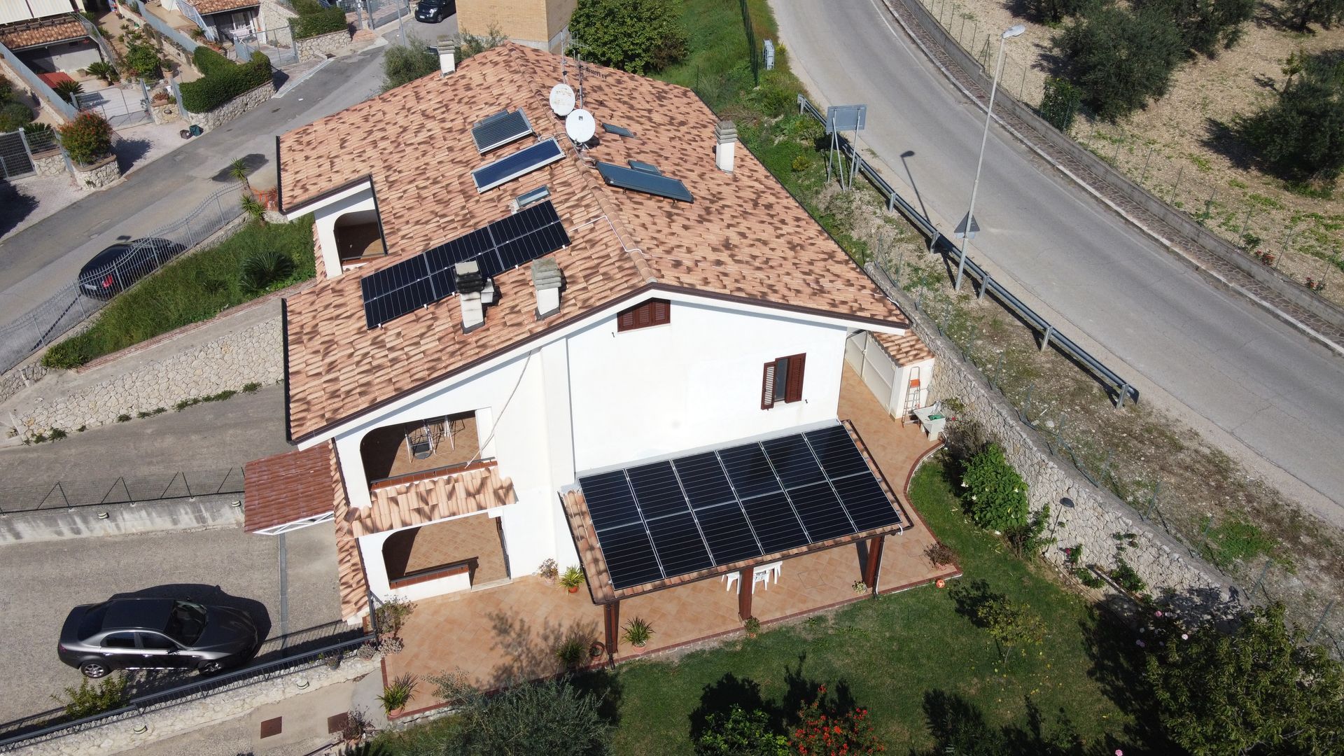 Veduta aerea di una casa bianca con tetto di tegole rosse, pannelli solari e un'auto parcheggiata accanto ad essa lungo una strada.