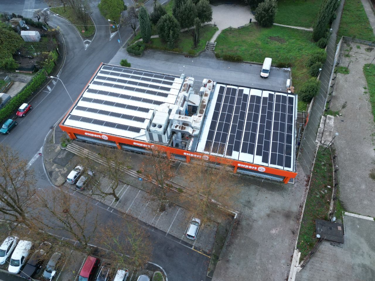 Veduta aerea di un grande edificio con tetto piano e pannelli solari, circondato da alberi e parcheggi.