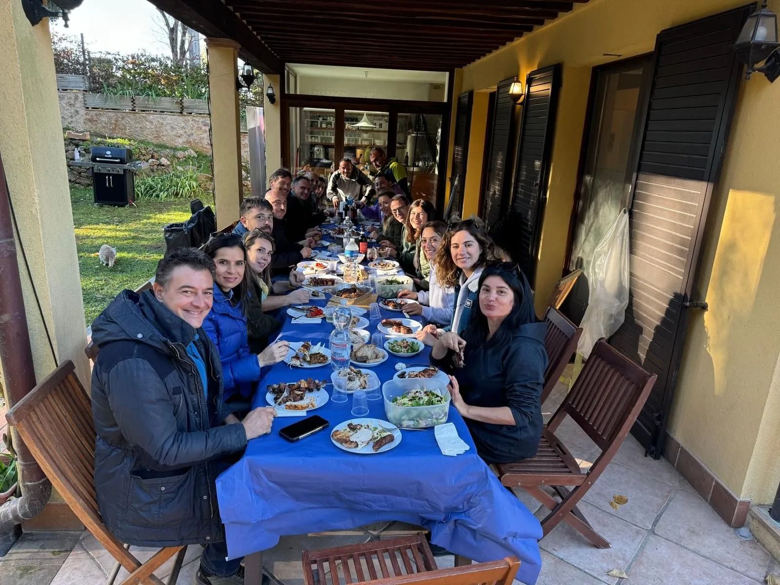 Gruppo di persone che pranzano attorno a un lungo tavolo coperto da una tovaglia blu in un patio esterno.