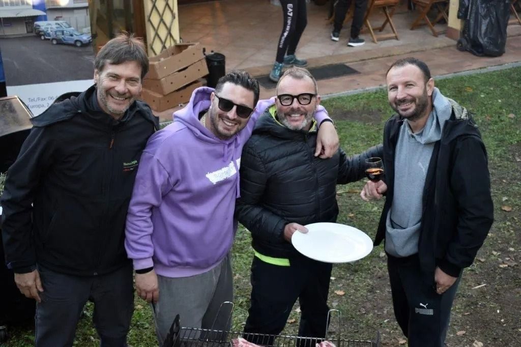 Quattro persone sorridenti all'aperto, una delle quali tiene in mano un piatto bianco, con indosso giacche casual e una felpa con cappuccio viola.