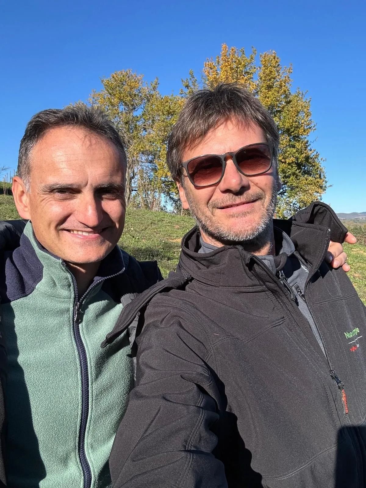 Due persone sorridenti all'aperto, sotto il sole, con alberi autunnali e un cielo azzurro limpido alle loro spalle.