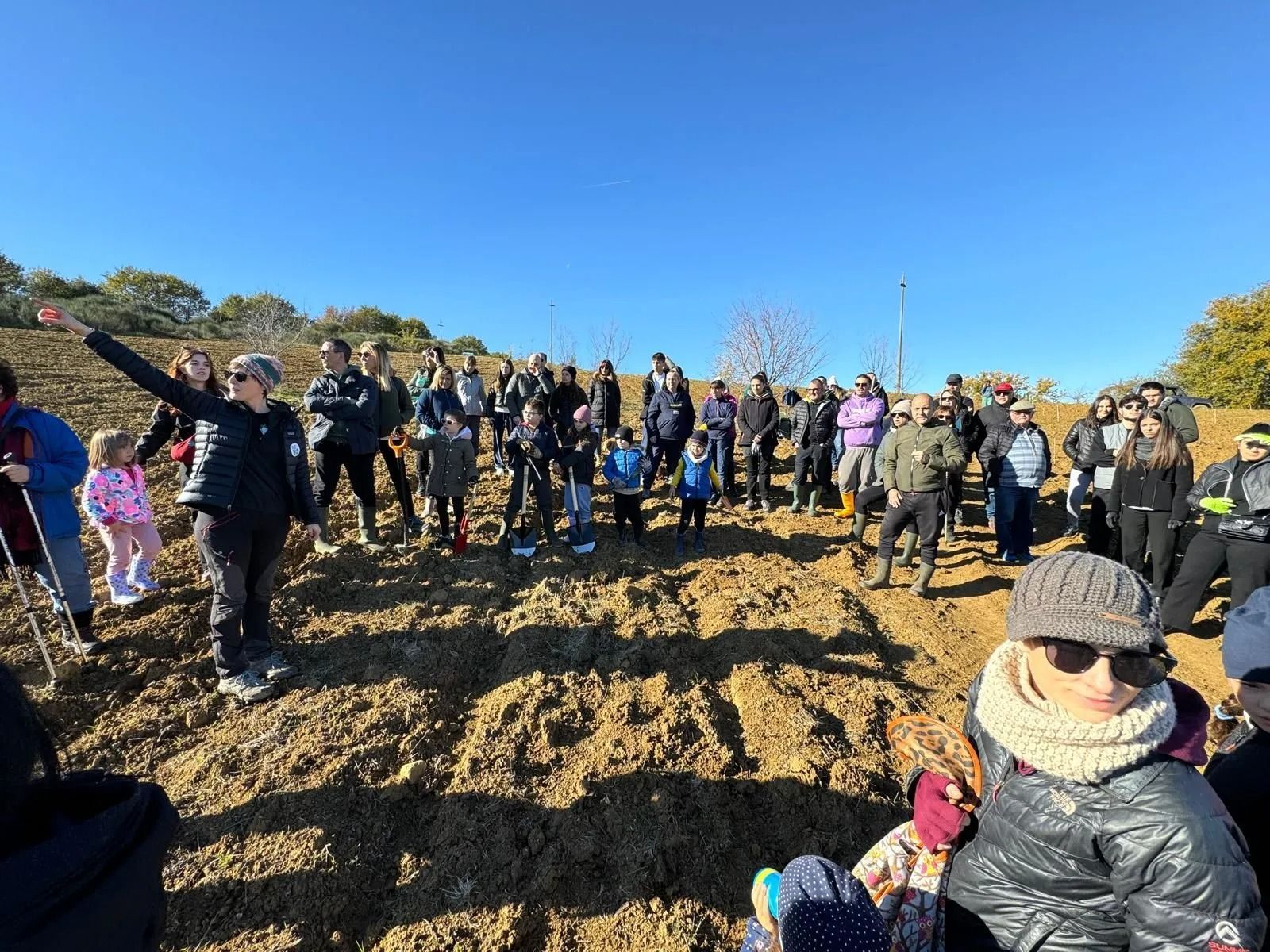 Una folla si è radunata all'aperto in un campo fangoso sotto un cielo azzurro e limpido; alcune persone salutavano con la mano e una era seduta in primo piano.