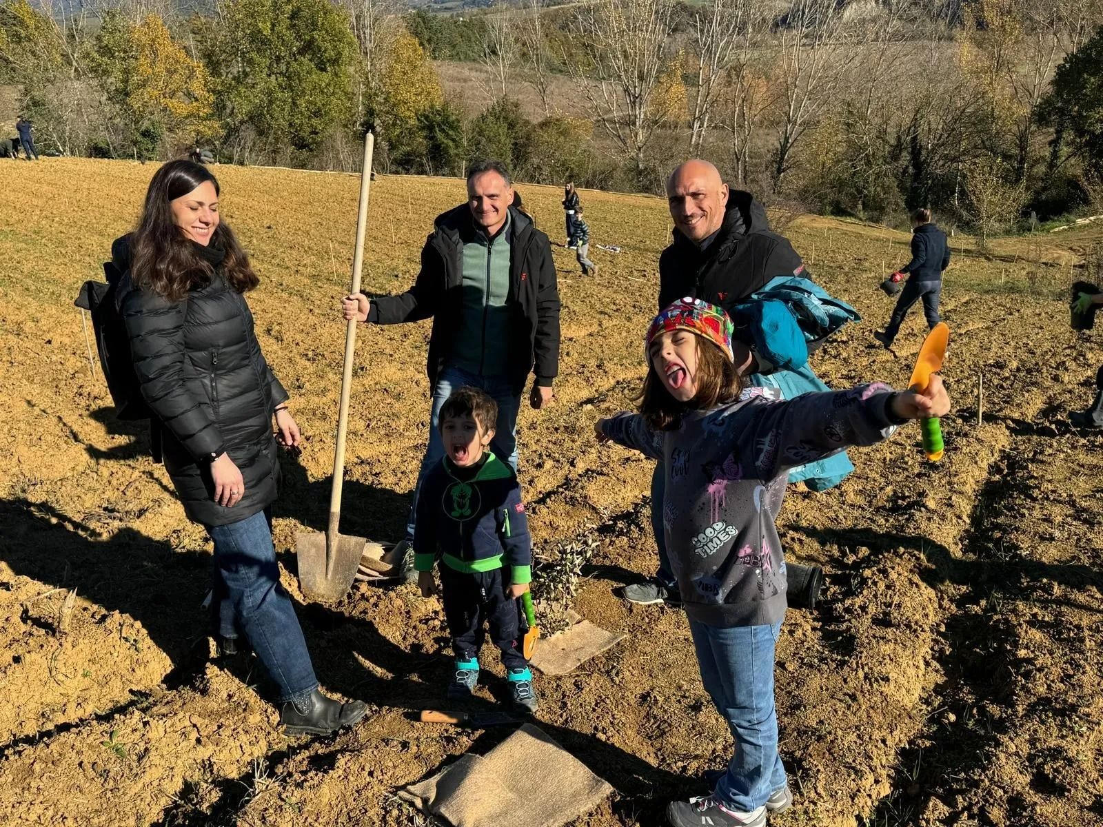 Persone in piedi in un campo fangoso con una pala, che reggono una piccola pianta in una giornata di sole.