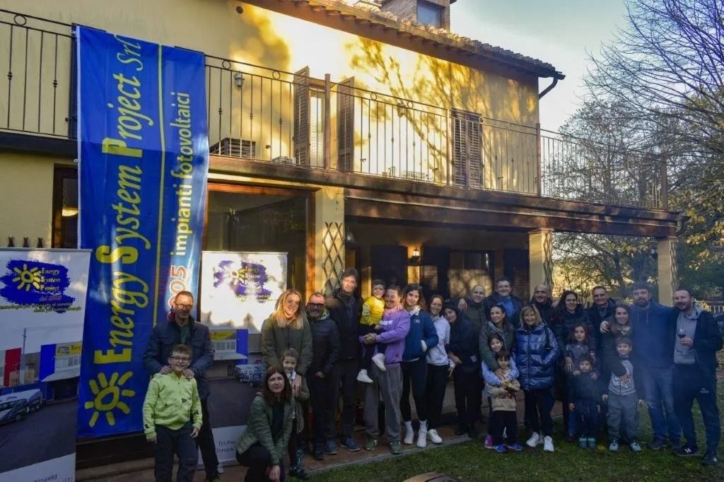 Gruppo di persone in posa davanti a un edificio a due piani illuminato dal sole, con uno striscione blu e alberi autunnali sullo sfondo.