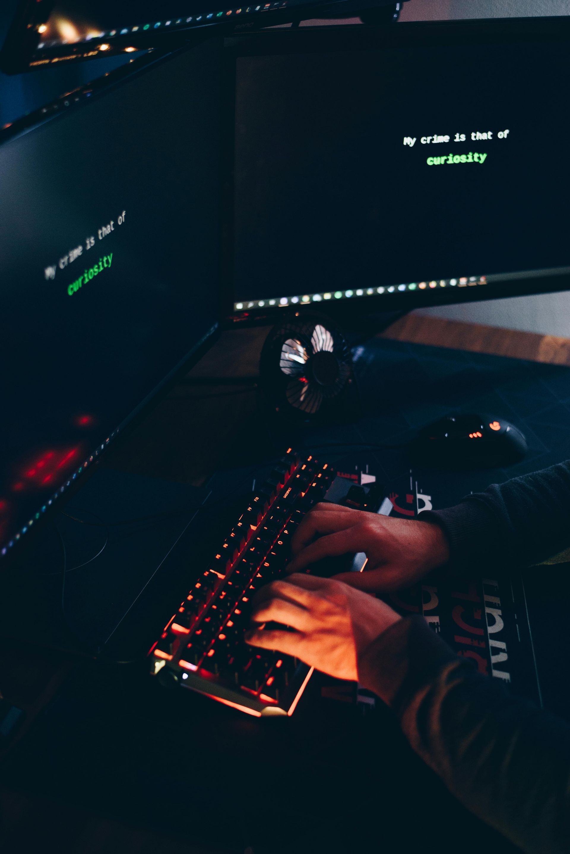 Hands typing on a lit-up keyboard in front of multiple computer monitors displaying code. Dark, indoor setting.