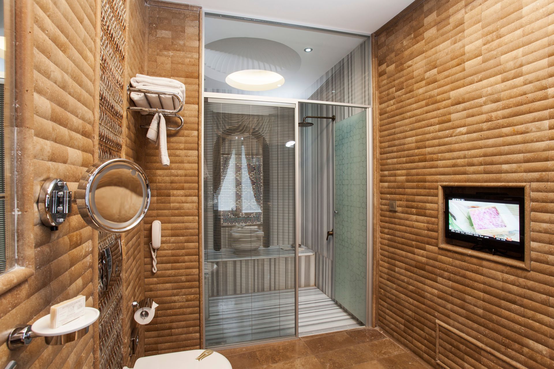Bathroom with brown brick-like tile walls. A shower, mirror, and small TV are visible.