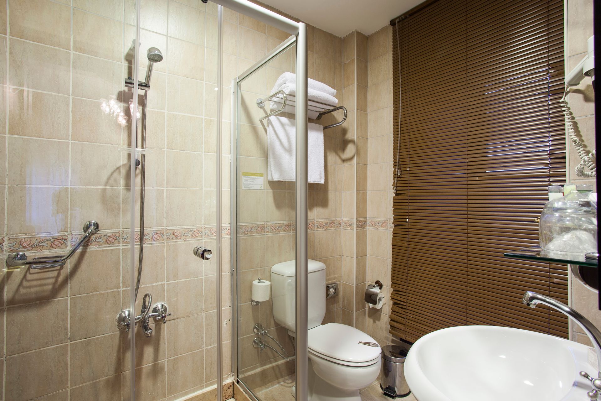 Ottoman Hotel Imperial| Small hotel bathroom with shower, toilet, and sink; beige tile walls; towels on rack.