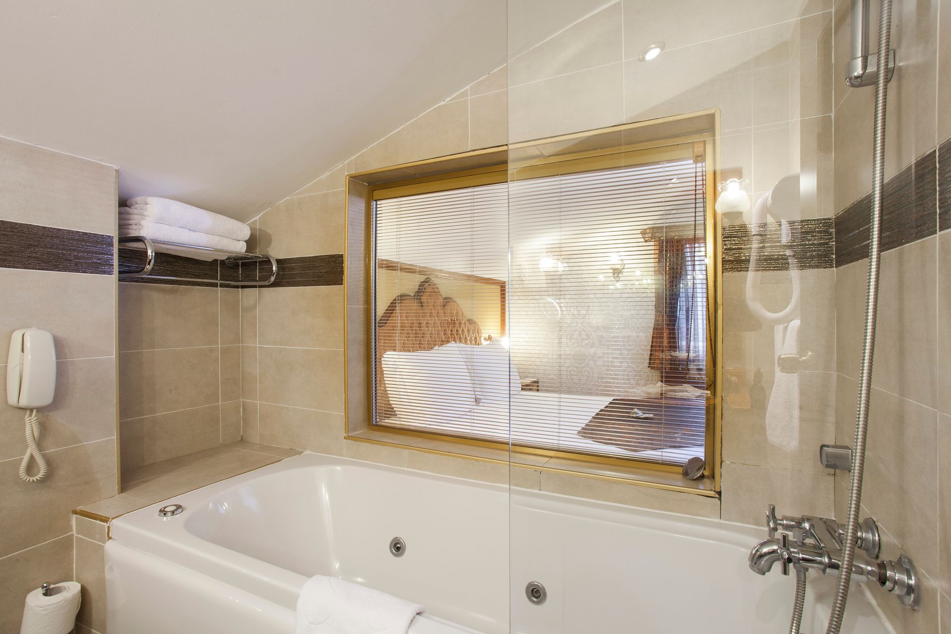 Bathroom with a white bathtub and shower. A window with a gold frame provides a view to the bedroom.
