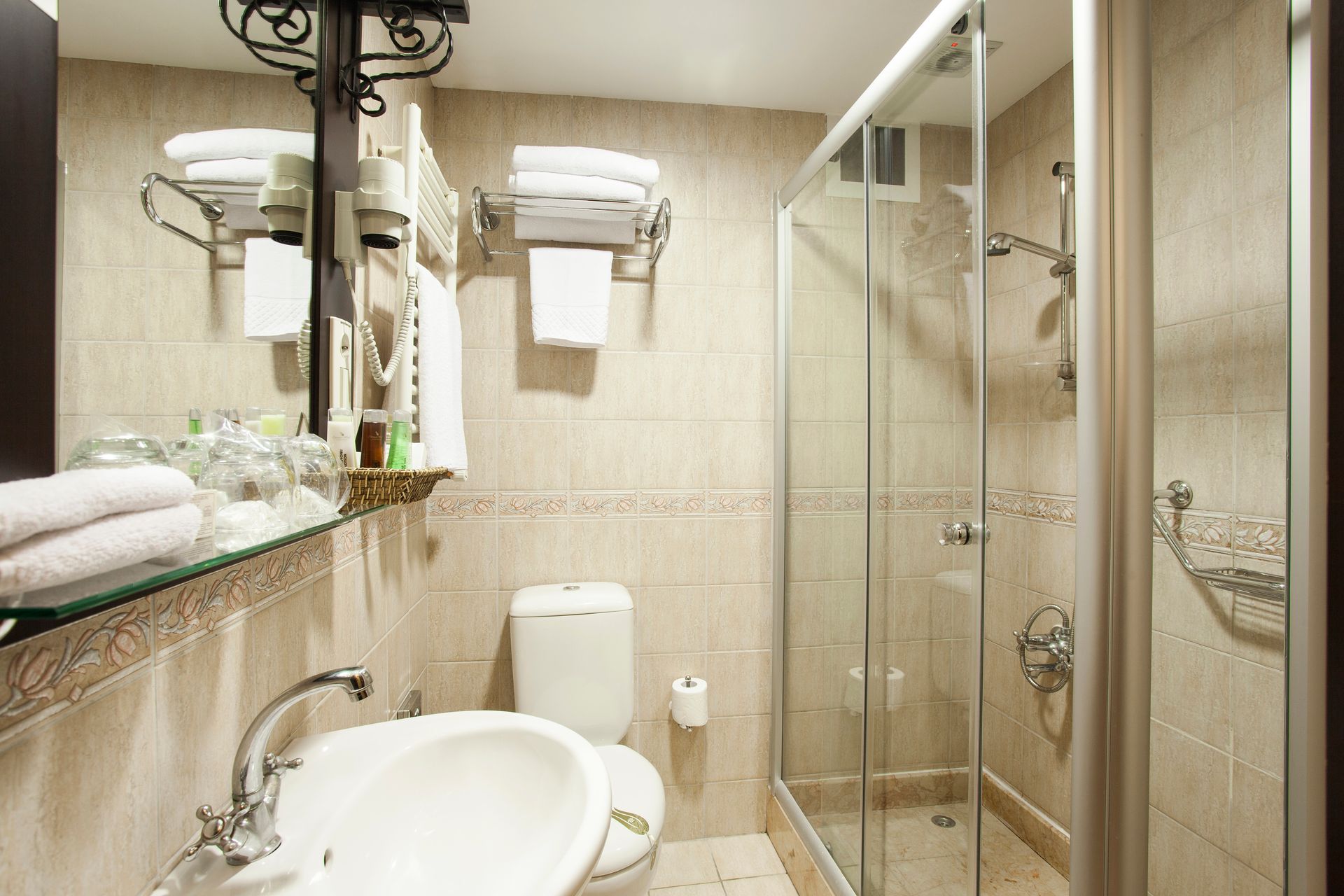 Bathroom with shower, toilet, sink, mirror, and towel rack. Beige tile walls.
