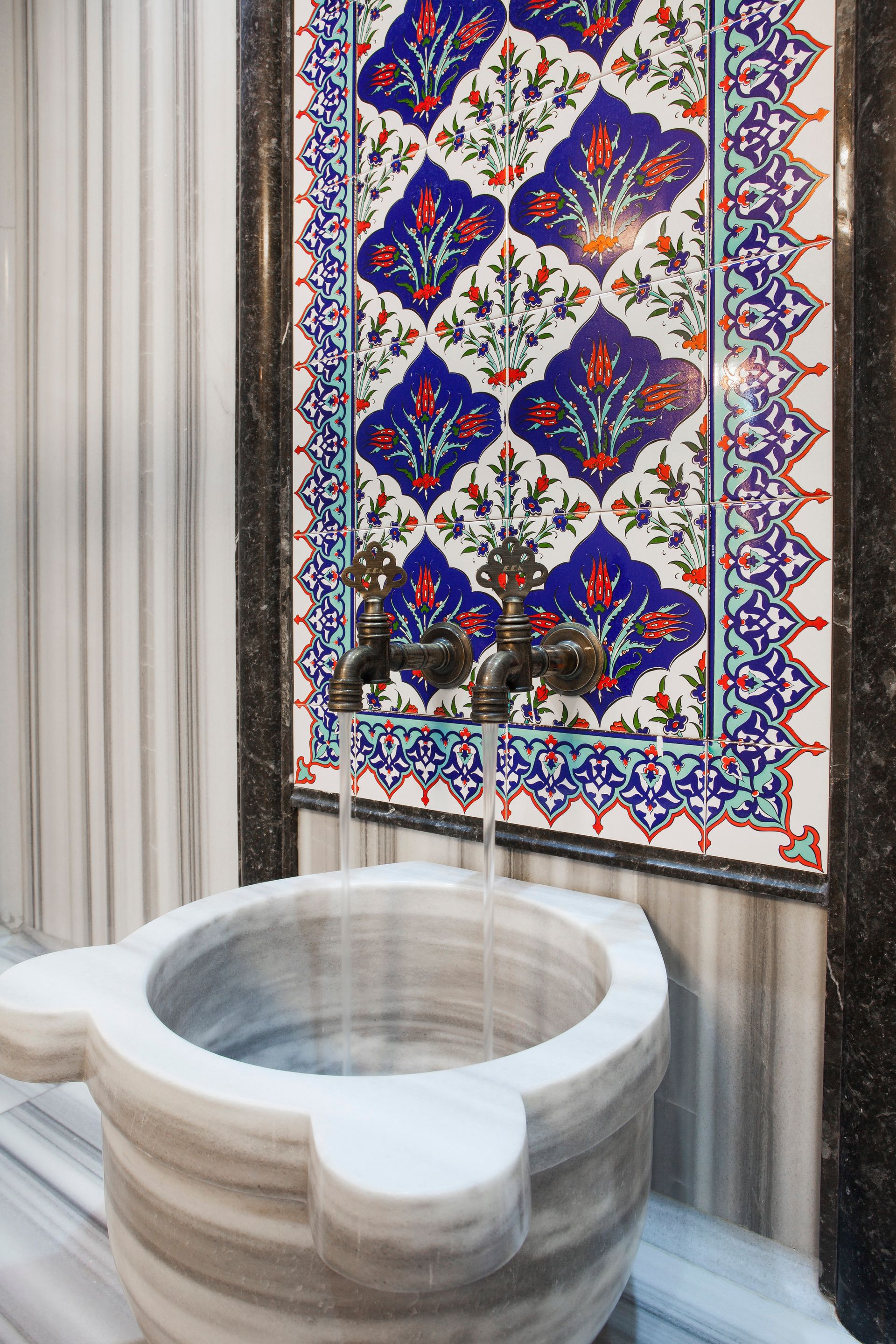 Water pouring from bronze taps into a marble basin, set against a colorful patterned tile wall.
