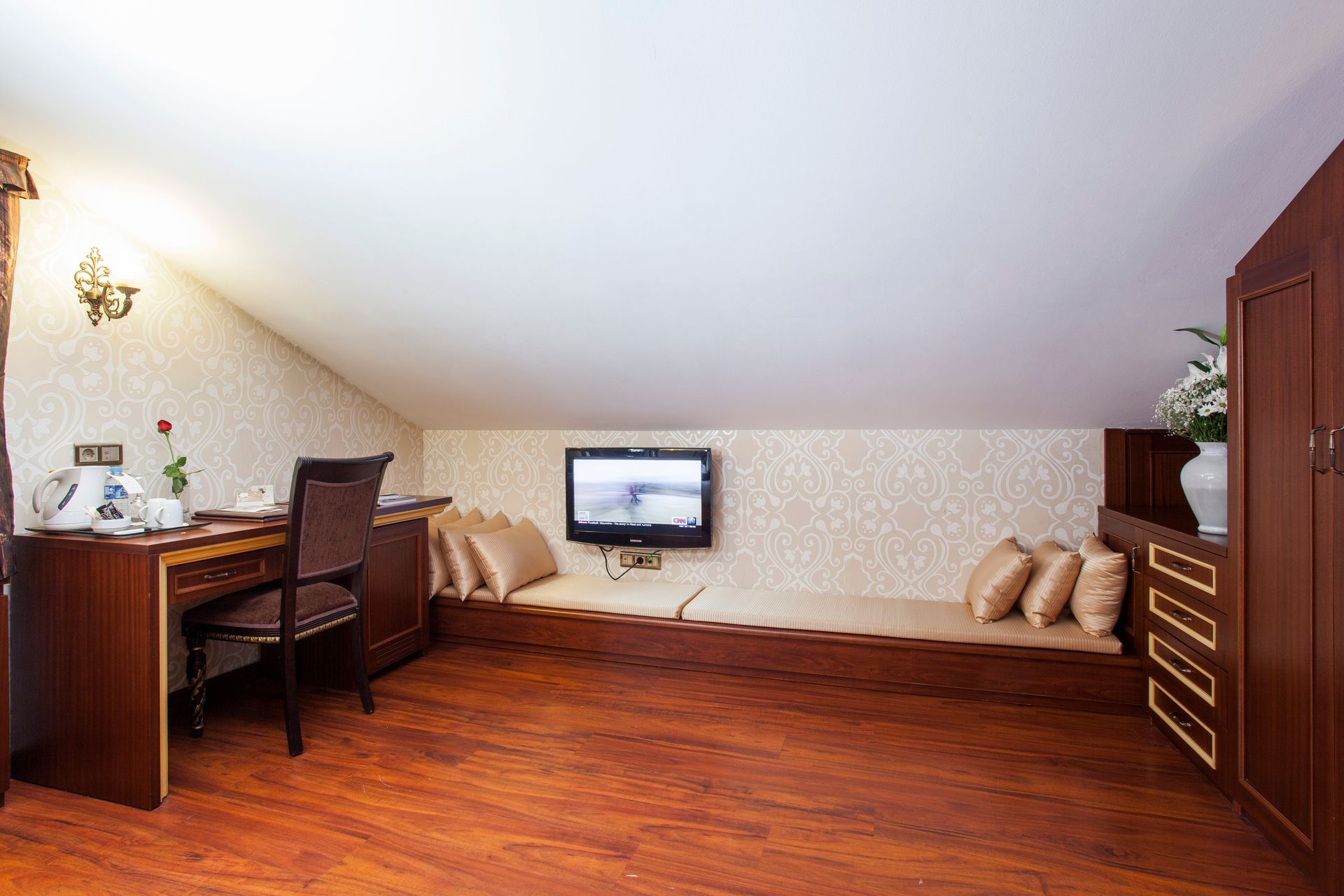 Attic room with wooden floor, desk, built-in sofa with cushions, TV, and cabinet.