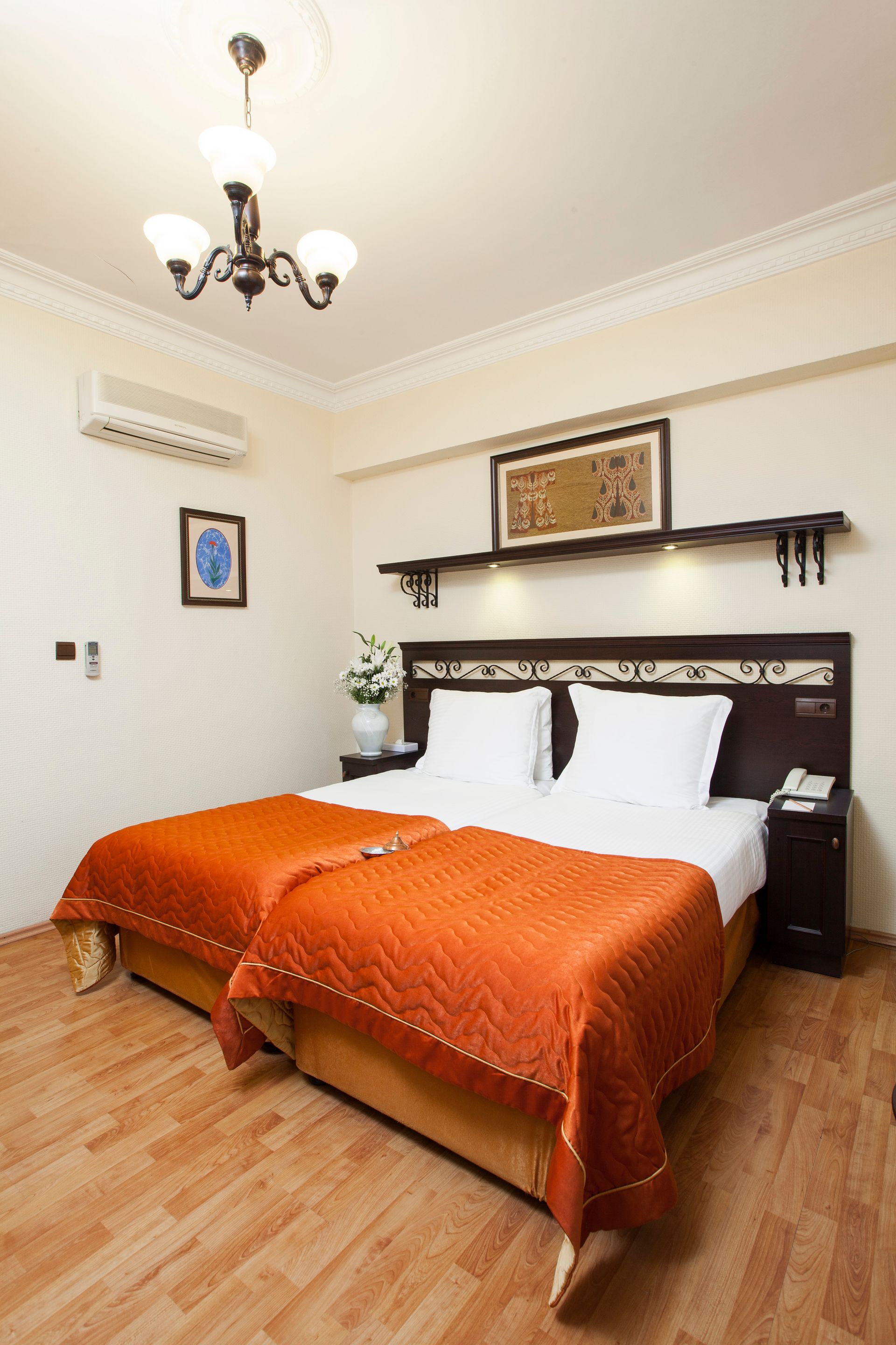 Hotel room with two beds, orange bedding, wooden floor, and a floral chandelier.