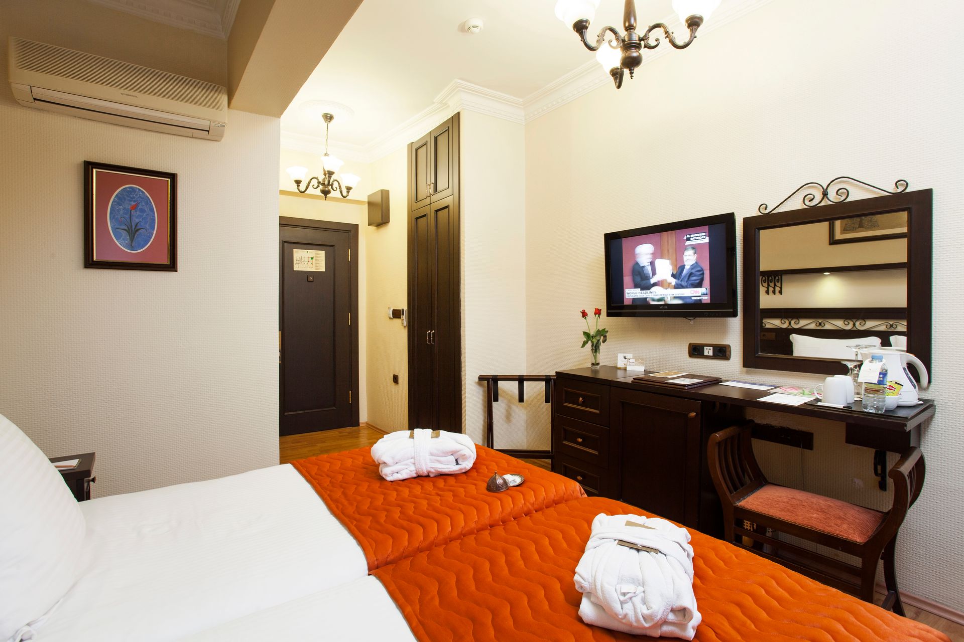 Hotel room with twin beds, TV, desk, and a door. Orange bedspreads, white walls, and a chandelier.