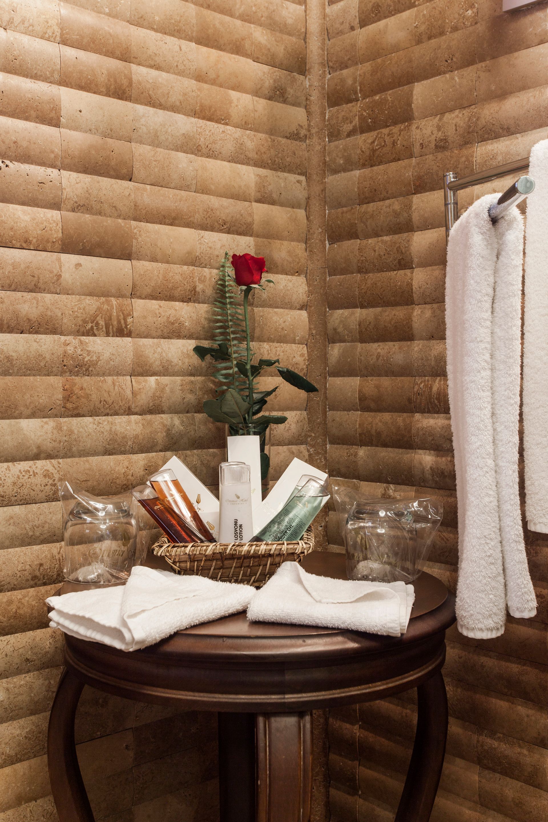 Bathroom corner with a round table holding toiletries, towels, and a rose.