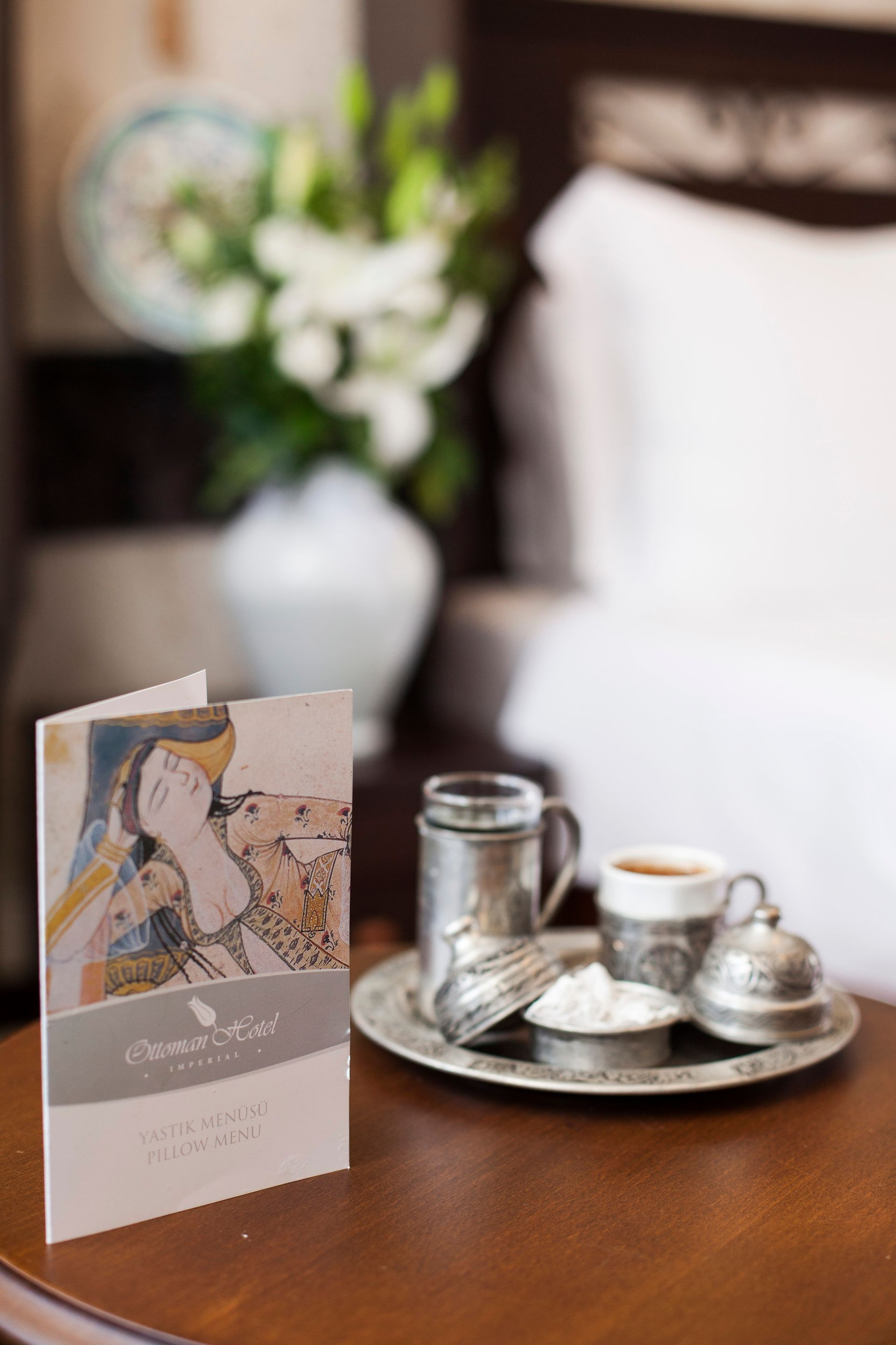 Close-up of Turkish coffee set and menu on a wooden table, with flowers and a bed in the background.
