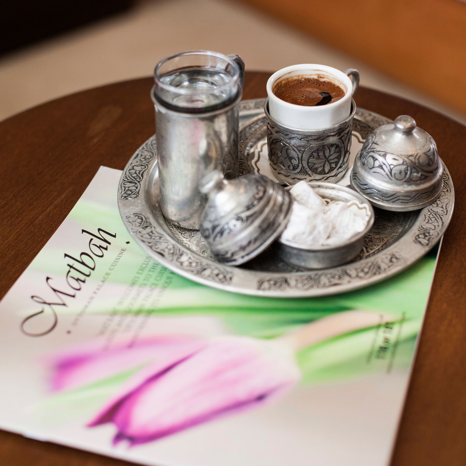 Turkish coffee setup on a silver tray with water, sugar, and a magazine on a wooden table.