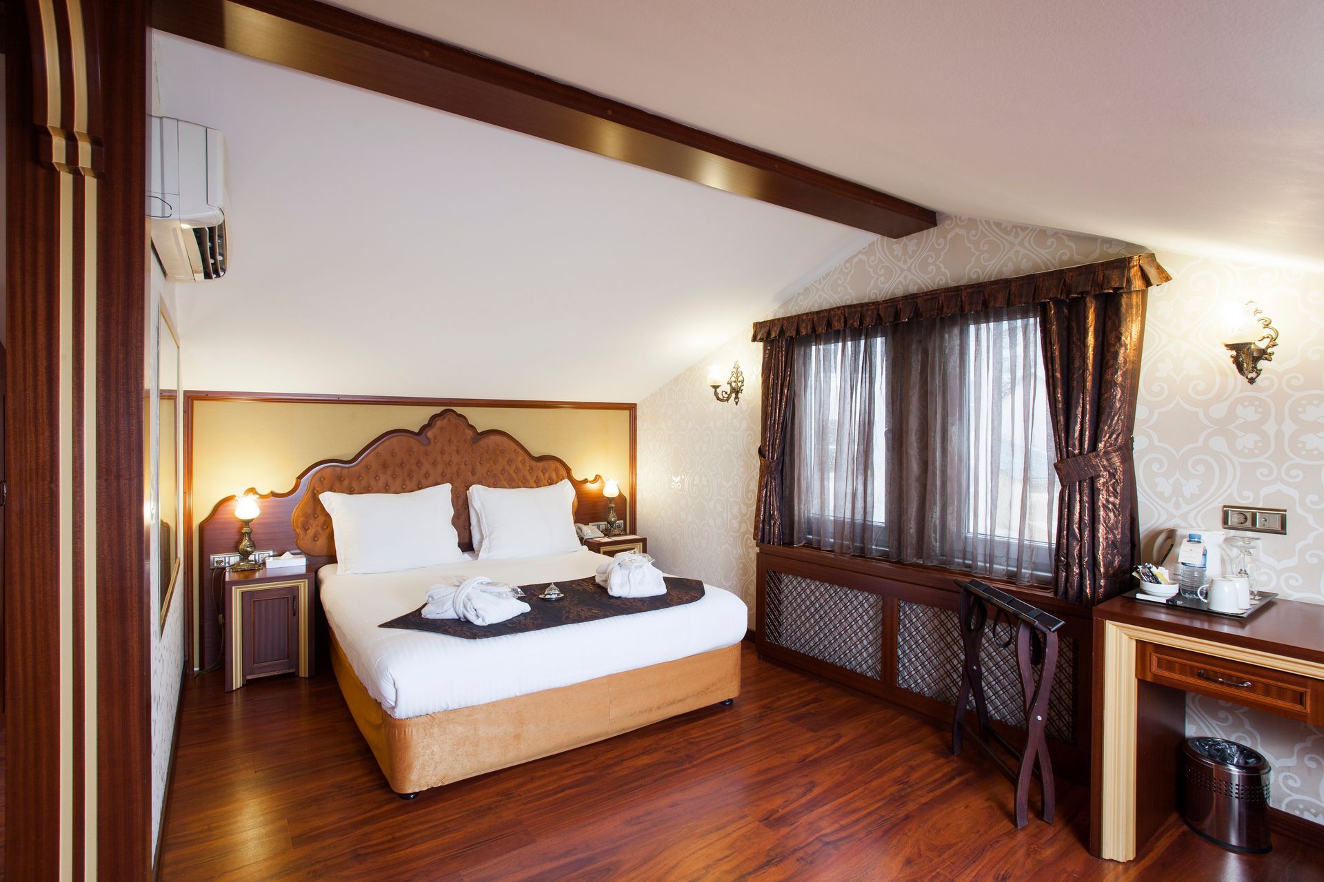 Hotel room with a bed, desk, and window with brown curtains; wooden floors and ceiling.