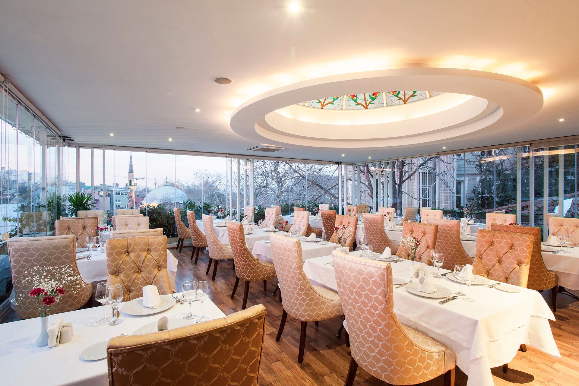 Restaurant dining room with tables set for guests, overlooking a daytime view. Beige chairs, white tablecloths.