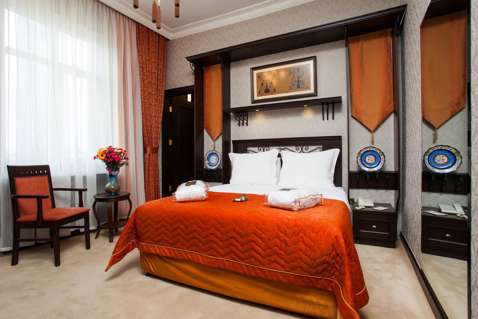 Ottoman Hotel, room with a queen bed featuring an orange quilted spread, wooden furniture, draped curtains, and decor plates.