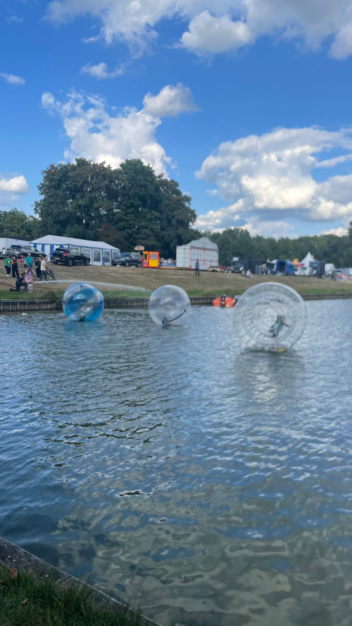 Mensen in doorzichtige opblaasballen op een meer met een park op de achtergrond. Helderblauwe lucht.