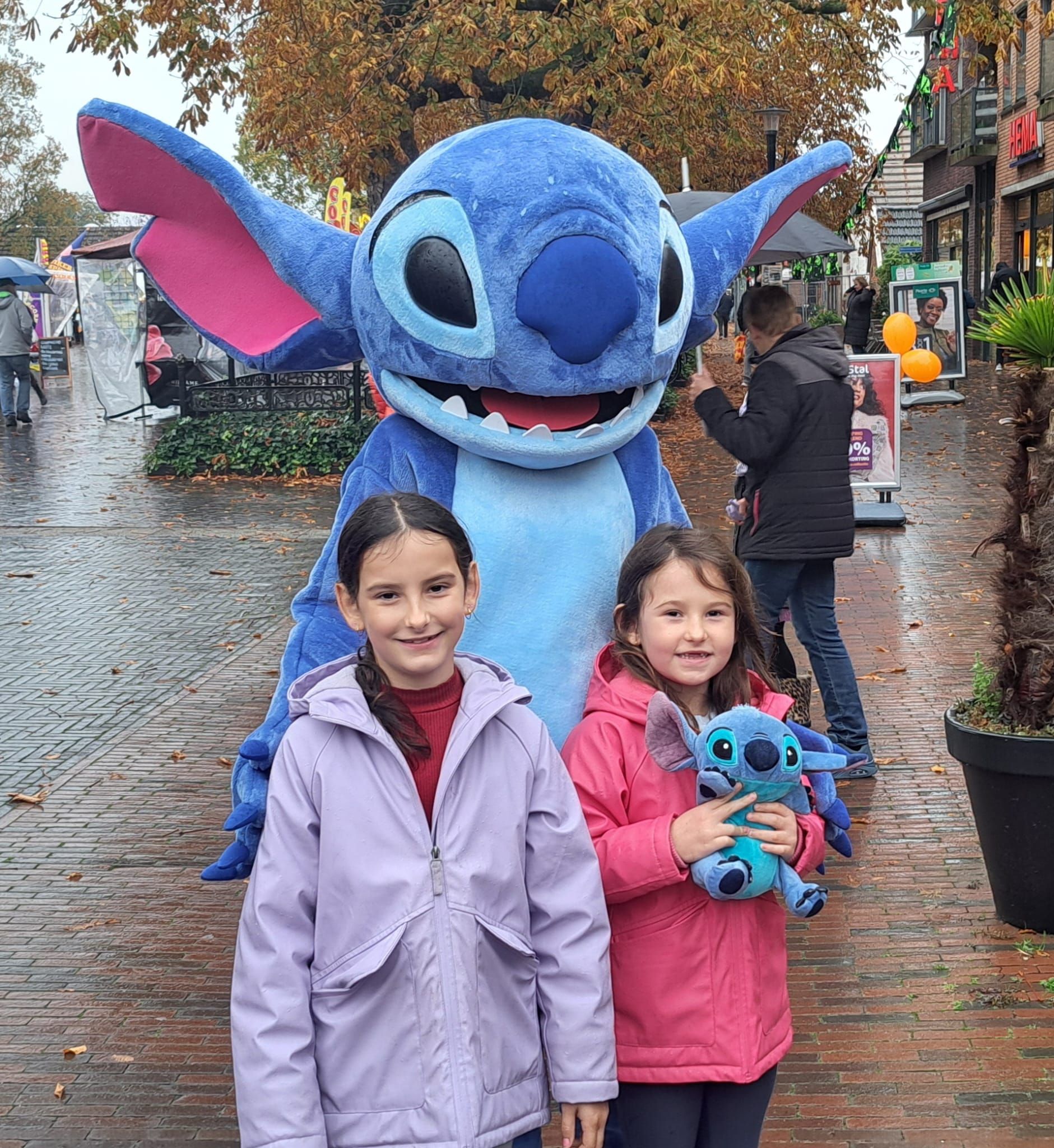 Twee meisjes poseren met een Stitch-figuur in een regenachtige buitenomgeving.