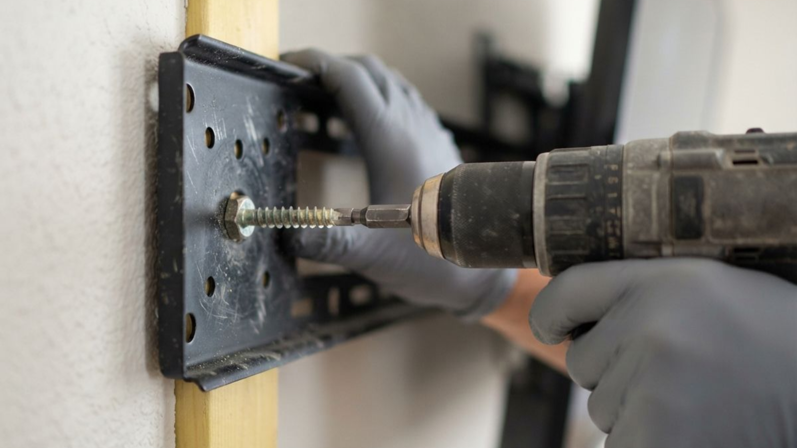 Gloved hands drilling a screw into a black wall bracket with a power drill