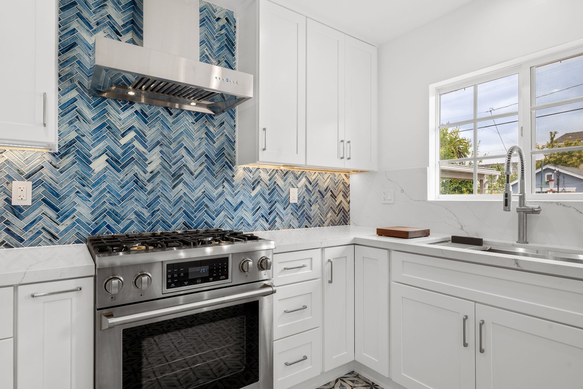 Bright white kitchen with blue herringbone backsplash, stainless stove, and sink by a window.