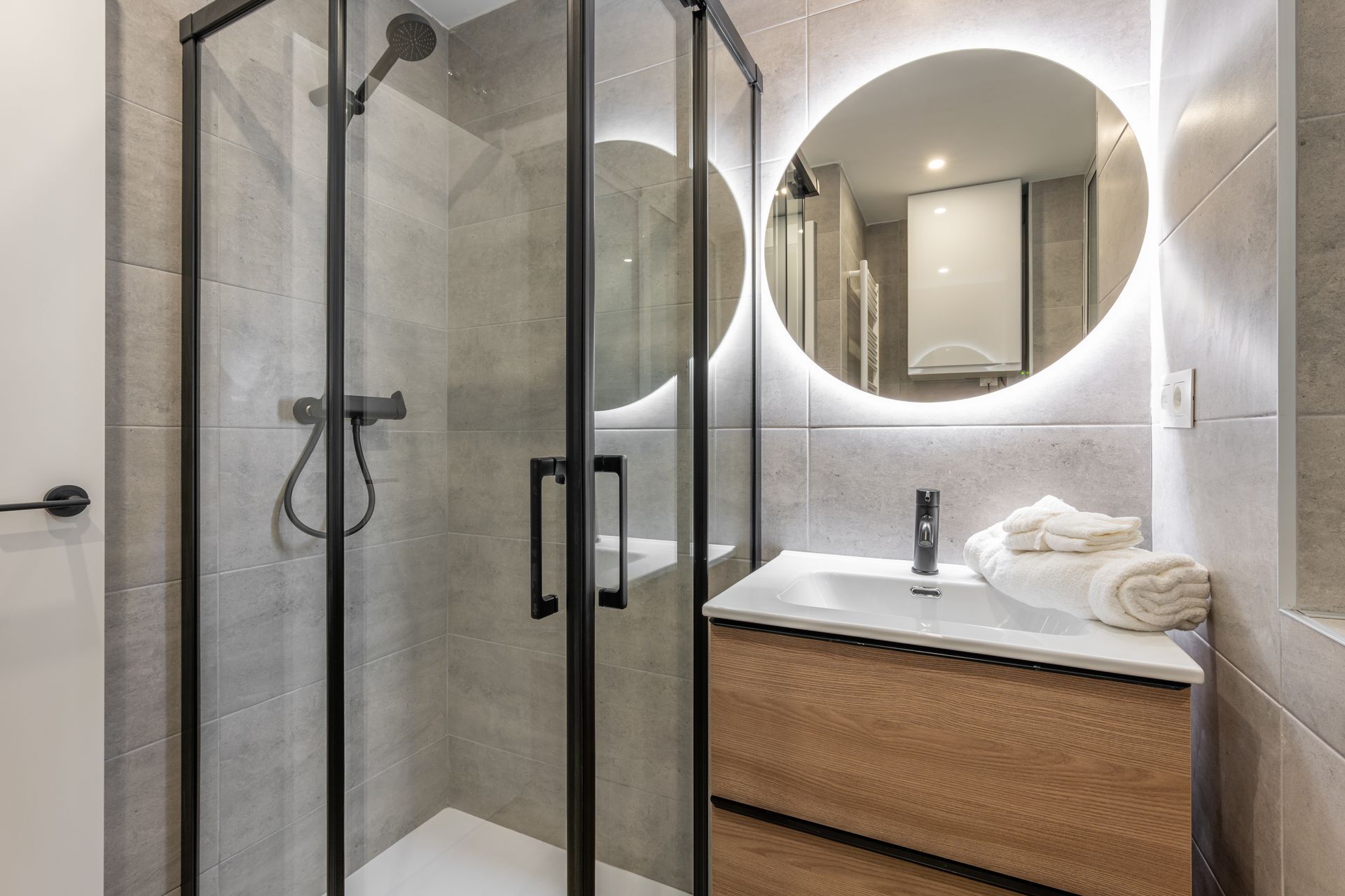 Bathroom with glass shower, wooden vanity, round backlit mirror. Gray tile walls.
