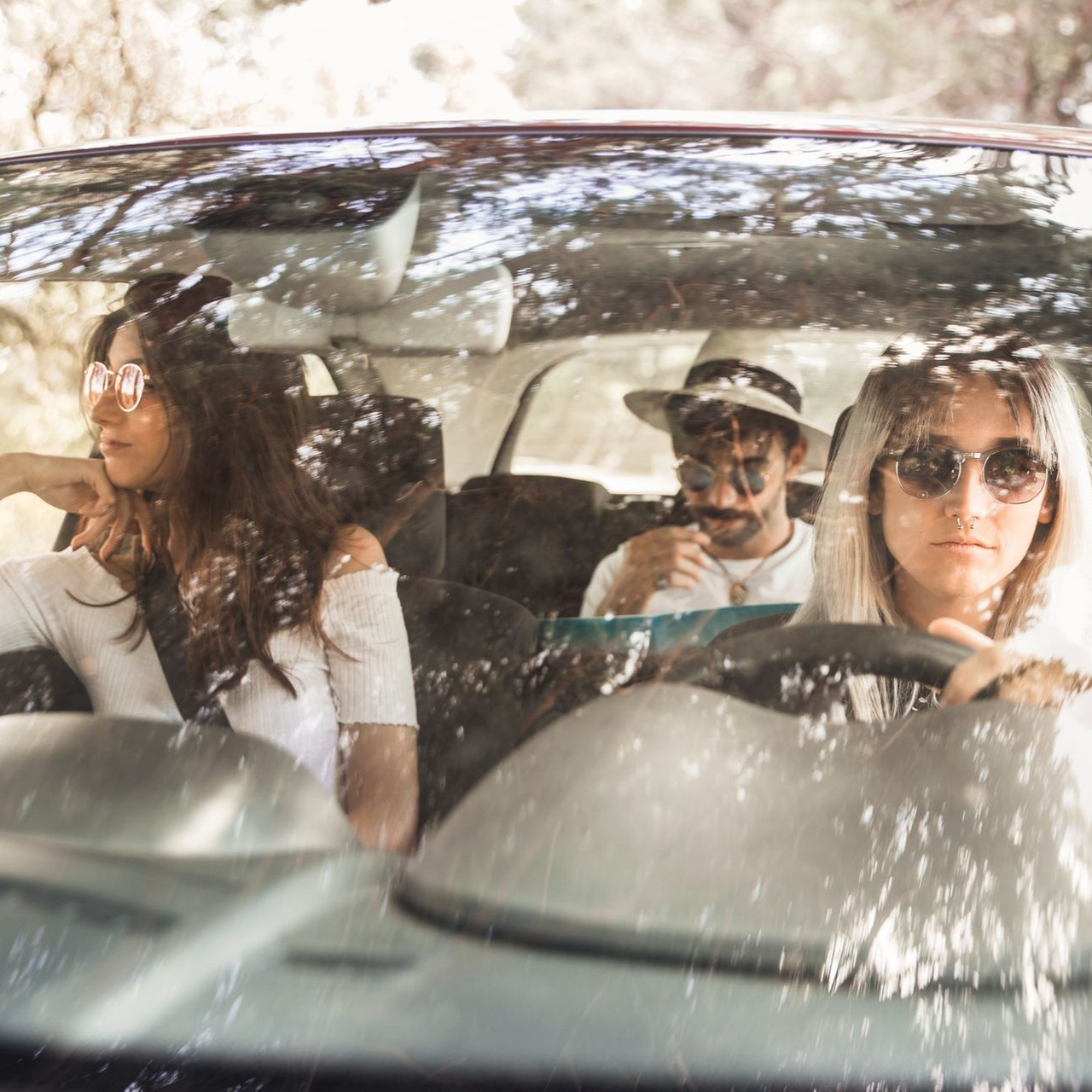 Three friends ride in a car, seen through the windshield with tree reflections.