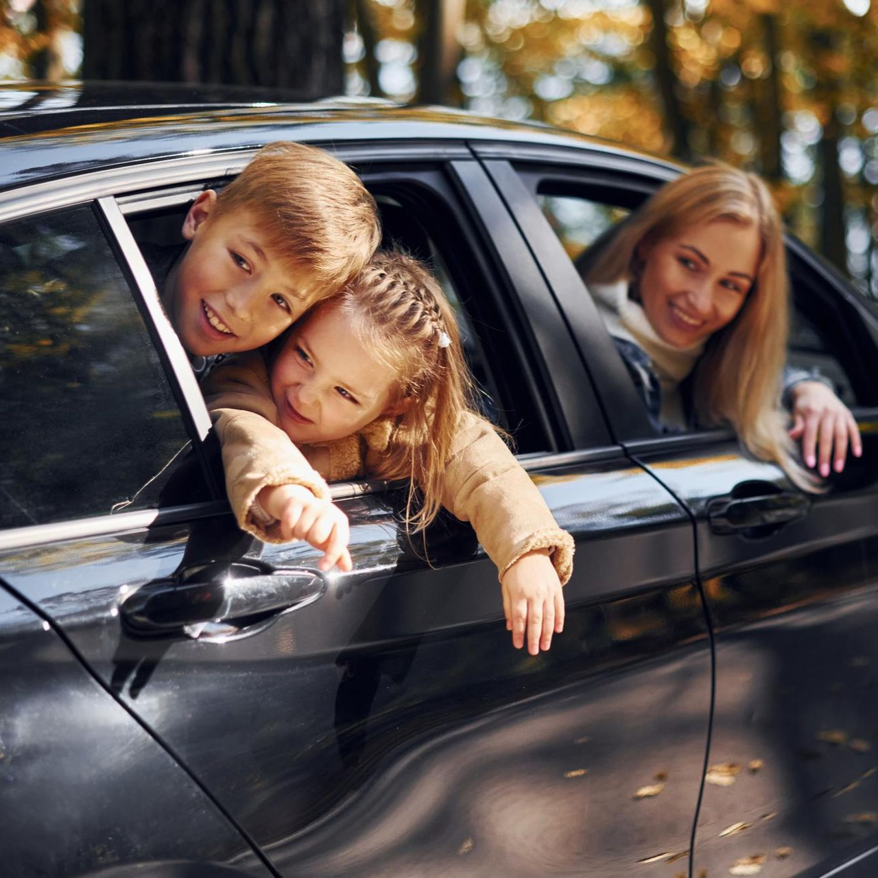 Two children lean out of a black car window while a woman smiles from the back seat.