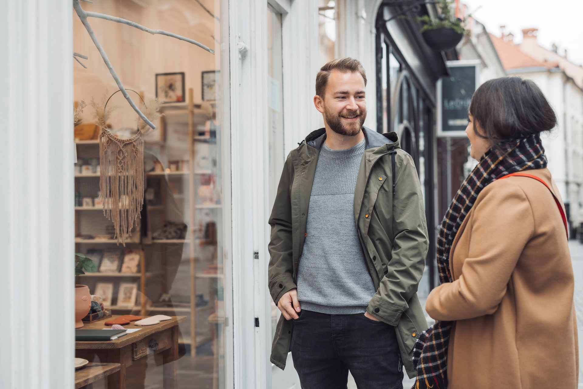 Two people chatting outside a storefront, one smiling in a green jacket and one in a tan coat with a scarf.