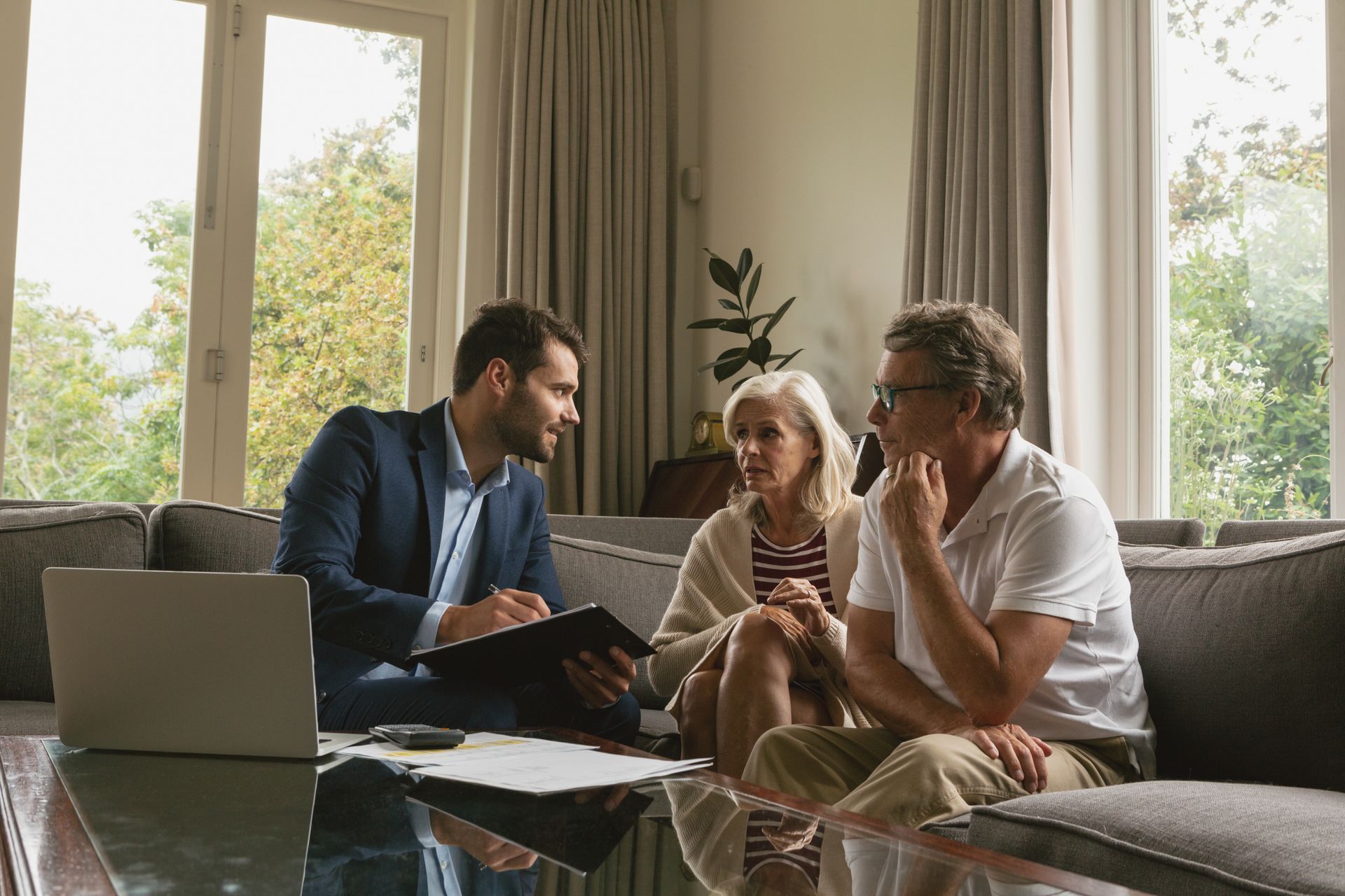 Three people discuss documents beside a laptop in a bright living room with large windows.