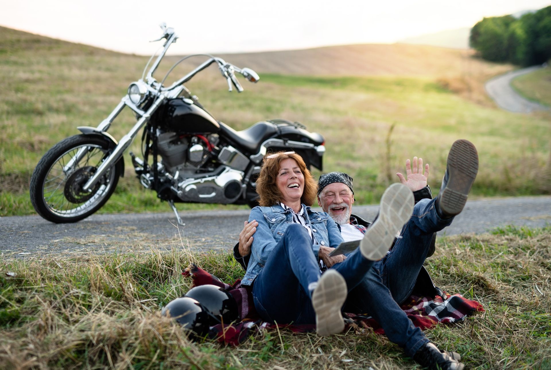 Two people laughing on grass beside a motorcycle, feet up in a sunny field.