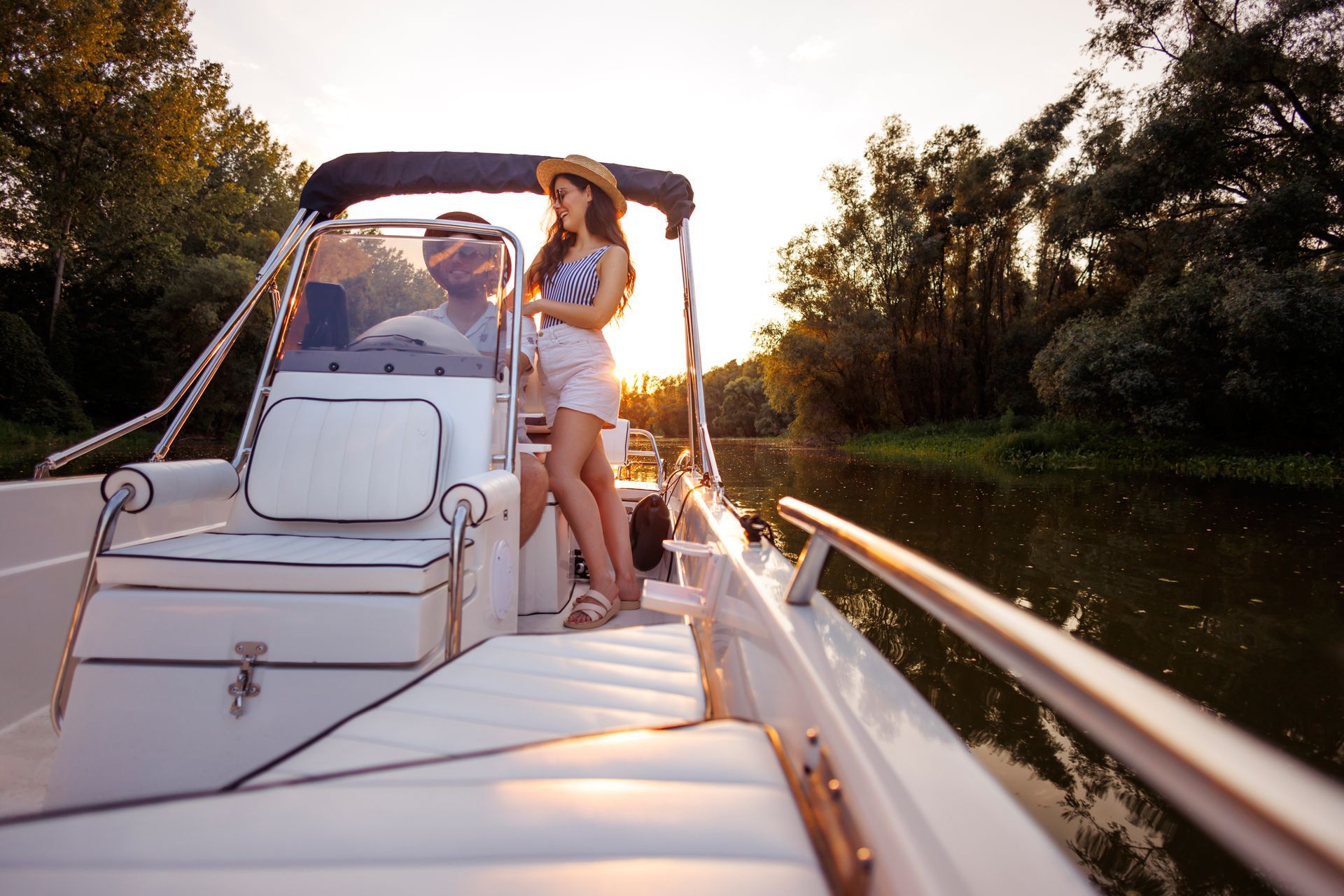 Two people on a motorboat at sunset, with one standing by the helm on a calm river.