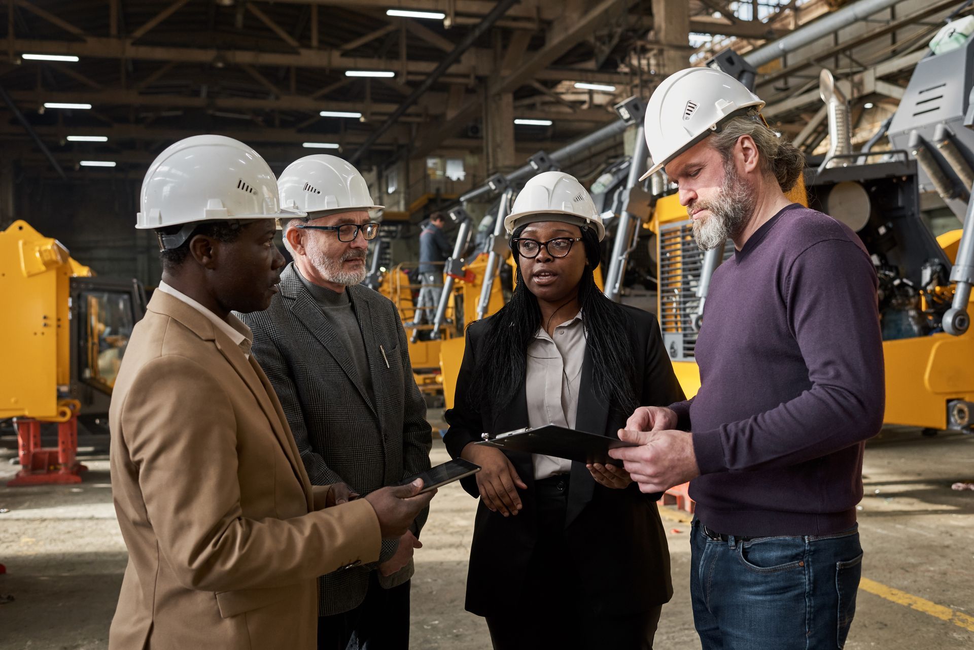 Instructor in blue shirt holding a hard hat and papers, speaking to workers in a warehouse.