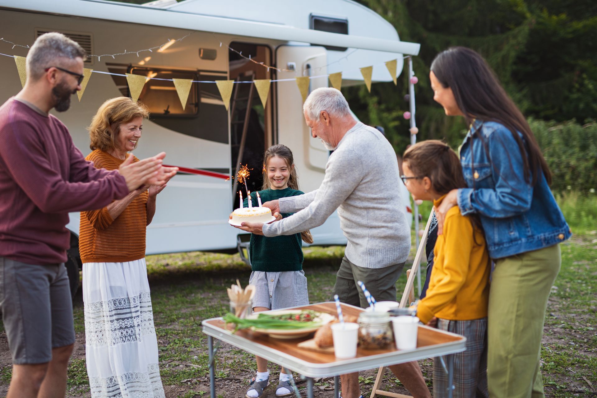 Family gathered beside a camper van, clapping and serving drinks at an outdoor picnic table.